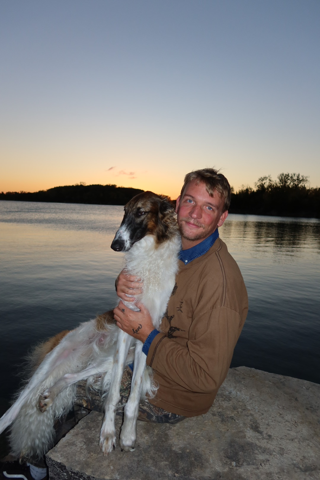 Maxwell Beck holding his dog beside the water at sunset